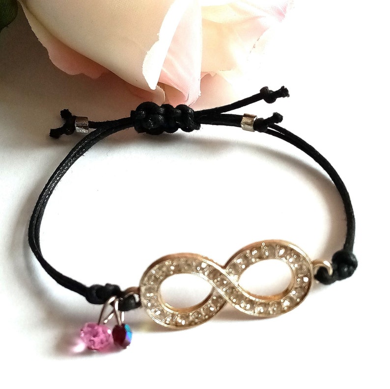 May include: Black string bracelet with an infinity symbol charm. The charm is silver and has clear rhinestones. The bracelet has two pink crystal charms hanging from it.