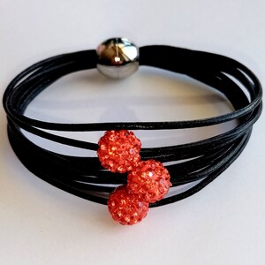 May include: A black leather bracelet with three red rhinestone balls. The bracelet is made of multiple strands of leather and has a magnetic clasp.