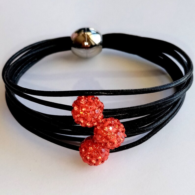 May include: A black leather bracelet with three red rhinestone balls. The bracelet is made of multiple strands of leather and has a magnetic clasp.