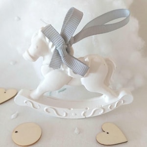May include: White ceramic rocking horse ornament with a grey satin ribbon bow. The ornament is hanging from a loop of ribbon.