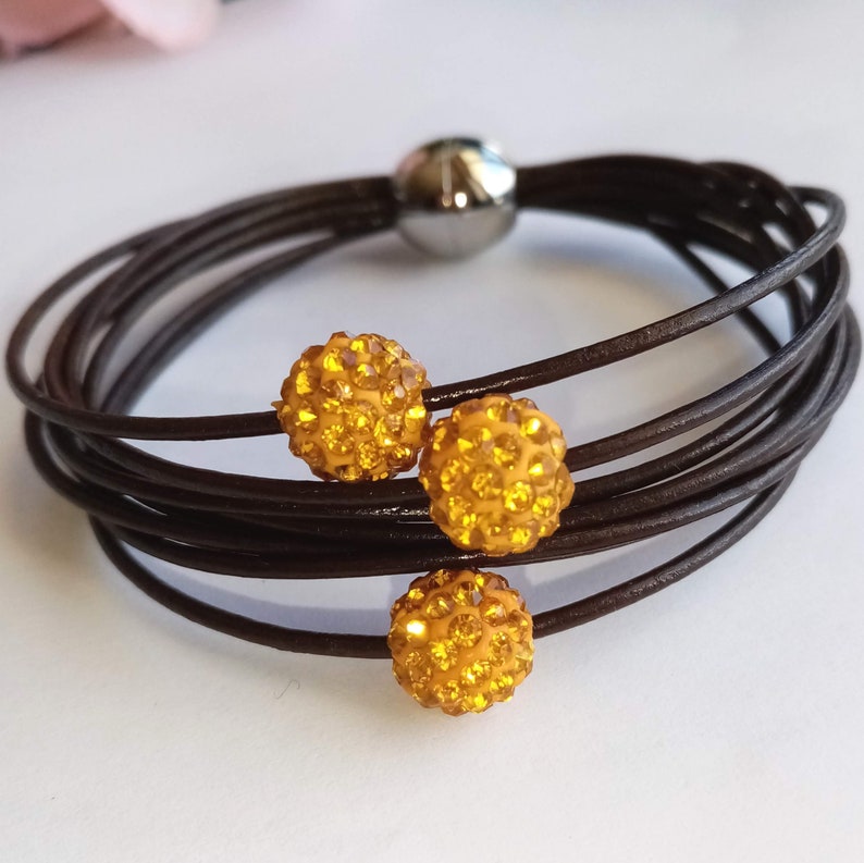 May include: A brown leather bracelet with three gold-toned, sparkly, round charms. The bracelet is made of multiple strands of leather.