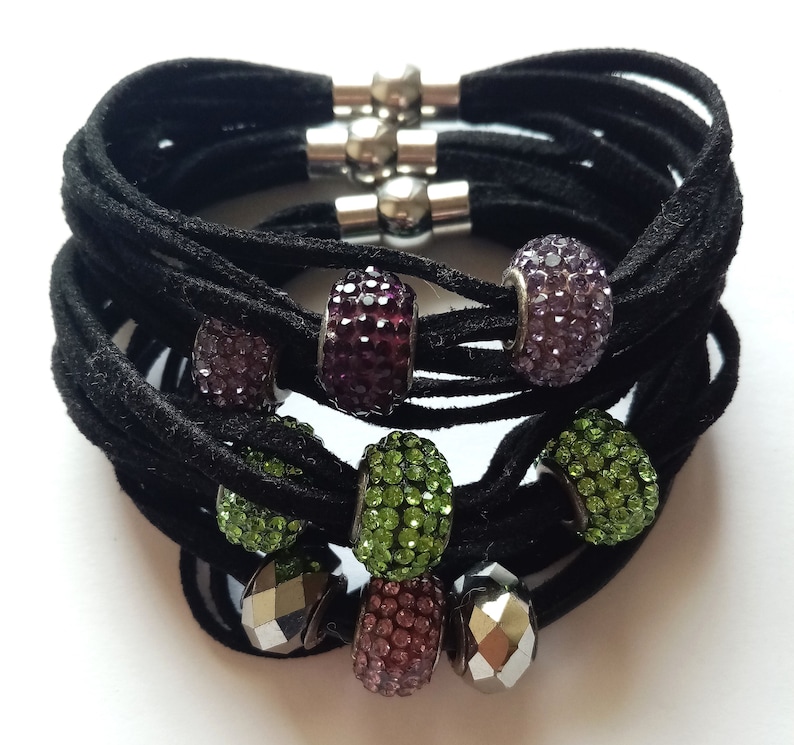 May include: A black suede bracelet with multiple strands and silver magnetic clasps. The bracelet is adorned with colorful crystal beads in shades of purple, green, and pink.