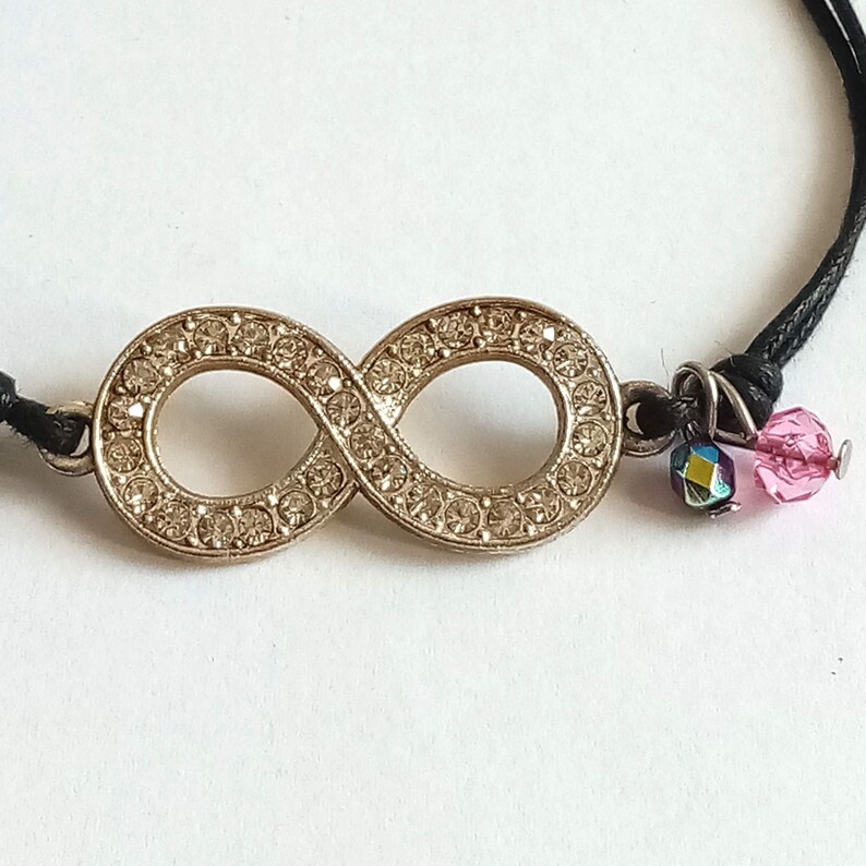 May include: A black cord bracelet with a gold infinity symbol charm encrusted with rhinestones and pink and blue crystal accents.