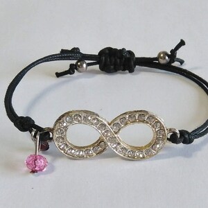 May include: A black string bracelet with an infinity symbol charm. The charm is silver and has clear rhinestones. A pink crystal dangles from the bottom of the bracelet.