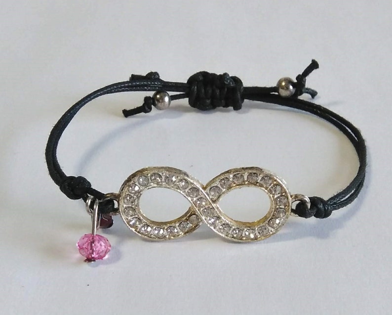 May include: A black string bracelet with an infinity symbol charm. The charm is silver and has clear rhinestones. A pink crystal dangles from the bottom of the bracelet.
