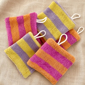 Puede incluir: Un conjunto de cuatro paños de lavado cuadrados de tela de rizo con rayas horizontales de colores vibrantes en tonos naranja, magenta, amarillo y morado. Cada paño incluye un pequeño lazo de tela blanca sujeto a una de las esquinas.