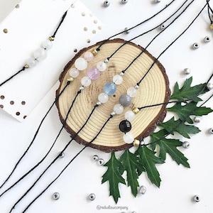 May include: Four black string bracelets with different coloured gemstone beads. The bracelets are arranged on a wooden surface with green leaves and silver beads scattered around. The bracelets feature white, pink, blue, grey, and black beads.
