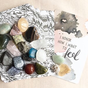 May include: A card with the text "I honor how I want to feel" in black calligraphy on a watercolor background with gray and white stars. A second card with a black and white illustration of leaves and branches is scattered with various colorful stones and crystals.