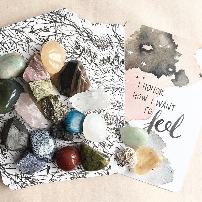 May include: A card with the text "I honor how I want to feel" in black calligraphy on a watercolor background with gray and white stars. A second card with a black and white illustration of leaves and branches is scattered with various colorful stones and crystals.