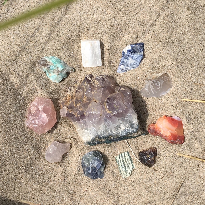 May include: A collection of crystals arranged in a circle on a sandy surface. The crystals include amethyst, rose quartz, clear quartz, carnelian, and a green stone.