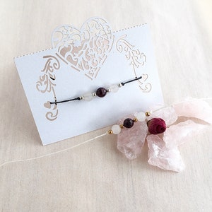 Peut inclure: Un bracelet délicat avec des perles blanches et rouge foncé sur un cordon noir, présenté sur une carte blanche avec un motif de cœur découpé. Le bracelet est accompagné d'une petite rose et de cristaux roses.
