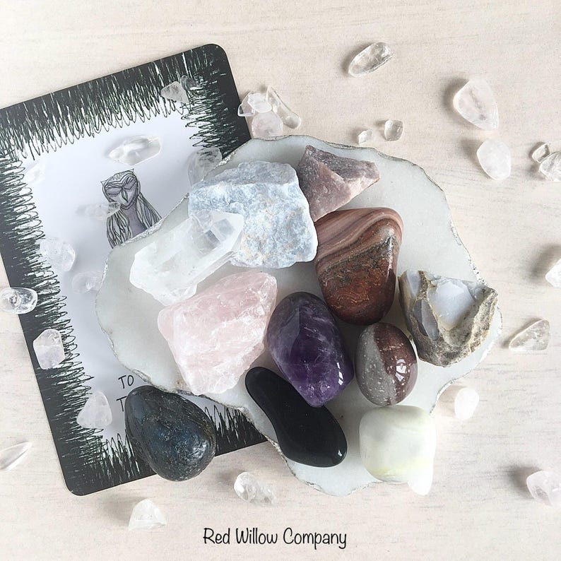 May include: A collection of 11 polished crystals and stones on a white surface. The crystals include amethyst, rose quartz, clear quartz, black obsidian, and various shades of brown and grey.