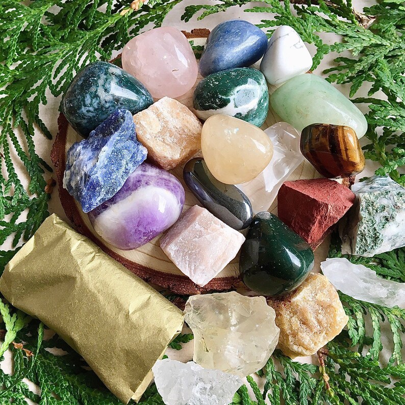 May include: A collection of various colored and textured stones, including amethyst, rose quartz, agate, and jasper. The stones are arranged on a wooden surface with a brown paper wrapping in the foreground.