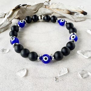 May include: A stretch bracelet with matte black onyx beads, interspersed with blue evil eye beads and silver-toned accents. The bracelet is designed for easy wear and is a stylish accessory.