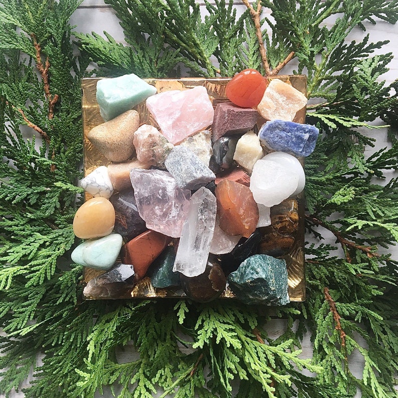 May include: A collection of colorful crystals and gemstones in various shapes and sizes, displayed on a gold tray. The crystals include clear quartz, rose quartz, amethyst, tiger's eye, and blue lace agate.