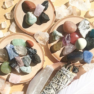 May include: A collection of crystals and stones on wooden coasters, including amethyst, rose quartz, clear quartz, and green aventurine. A bundle of sage is also included.