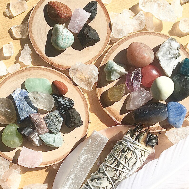 May include: A collection of crystals and stones on wooden coasters, including amethyst, rose quartz, clear quartz, and green aventurine. A bundle of sage is also included.