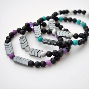 May include: Three beaded bracelets with black lava rock beads, gray chevron-shaped beads, and accent beads in turquoise, white, and purple. The bracelets are arranged on a white surface.