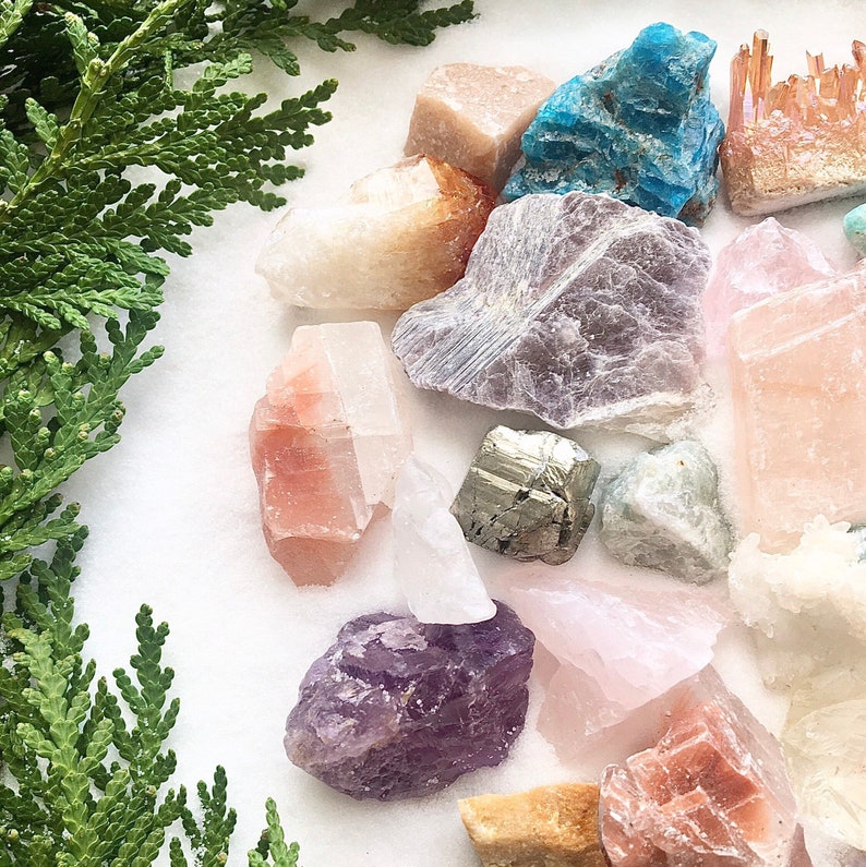 May include: A collection of various crystals and gemstones, including amethyst, rose quartz, clear quartz, pyrite, and blue apatite. The crystals are arranged on a white surface.