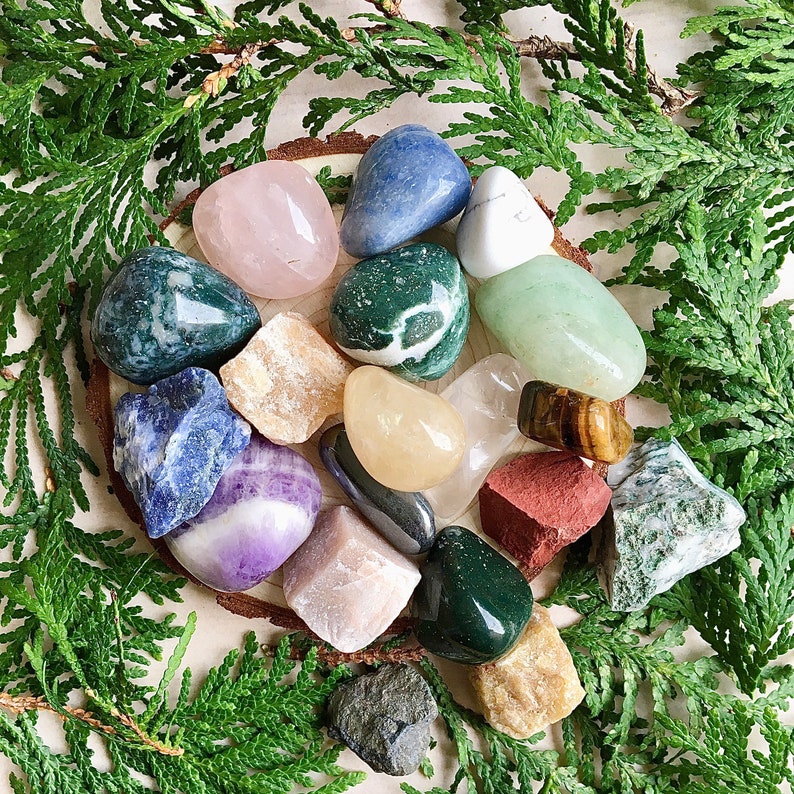 May include: A collection of 16 polished tumbled stones in various colors and shapes, including green, blue, pink, white, and brown. The stones are arranged on a light brown surface with green foliage surrounding them.