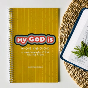 May include: A yellow workbook with the title "My God is" in red, white, and blue lettering. The subtitle is "WORKBOOK, a simple biography of God from the Psalms." The book has a spiral binding and is on a white surface.