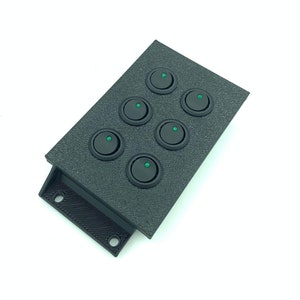 Op de afbeelding: Een zwart bedieningspaneel met zes ronde, zwarte schakelaars. Elke schakelaar heeft een klein groen lampje. Het paneel heeft een gestructureerd oppervlak en een montagebeugel met twee gaten.