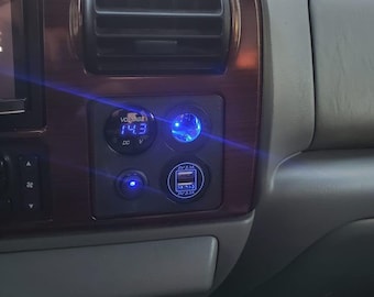 May include: A close-up of a car's dashboard with a digital voltage meter reading 14.3 volts, a blue LED light, and a dual USB charging port with a blue LED light.