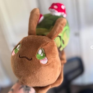 Puede incluir: Un juguete de peluche con forma de caracol con cuerpo marrón, ojos verdes grandes y un pequeño hongo en la espalda. El hongo tiene un gorro rojo con lunares blancos. El caracol tiene una cara sonriente y detalles rosas.