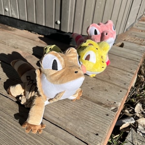 May include: Three plush toy lizards in different colours: brown, yellow and pink. The lizards are all smiling and have big eyes. They are sitting on a wooden surface.