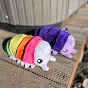 May include: Two plush toy isopods. One is rainbow-colored, with segments of yellow, orange, pink, and blue. The other is purple. Both have white faces, small black eyes, and tiny legs. They are resting on a weathered wooden surface.