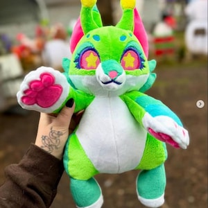 May include: A plush toy creature with a green body, white belly, and teal feet. It has pink ears, yellow antennae, and large eyes with star-shaped pupils. The creature's paw is pink and white.