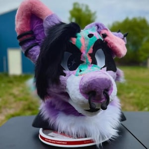 May include: A colorful furry head with pink, purple, black, and teal accents. The head features large eyes, a purple nose with a black ring, and a white fur trim. The ears are pink and purple. The head is sitting on a turntable.