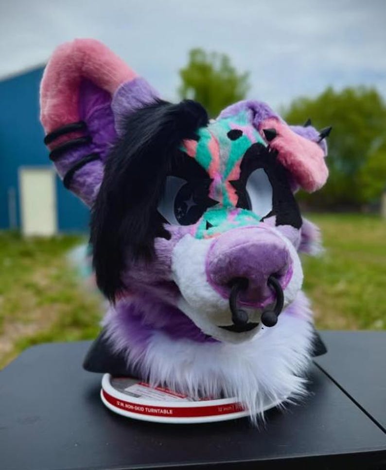 May include: A colorful furry head with pink, purple, black, and teal accents. The head features large eyes, a purple nose with a black ring, and a white fur trim. The ears are pink and purple. The head is sitting on a turntable.