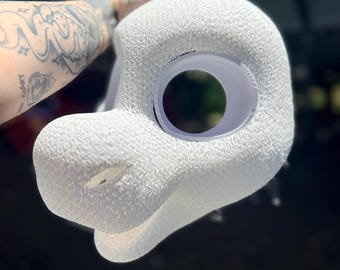 May include: A white, textured foam mask base featuring a rounded head shape, a protruding snout, and a large, circular eye opening. The surface has a porous, granular appearance, and the structure includes a defined jawline and a small nose opening.