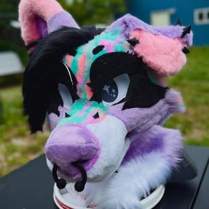 May include: A colorful fursuit head featuring a purple, pink, black, and teal design. The furry head has large, expressive eyes, a purple nose with a black ring, and black spikes on the ears. The fursuit is resting on a white surface.