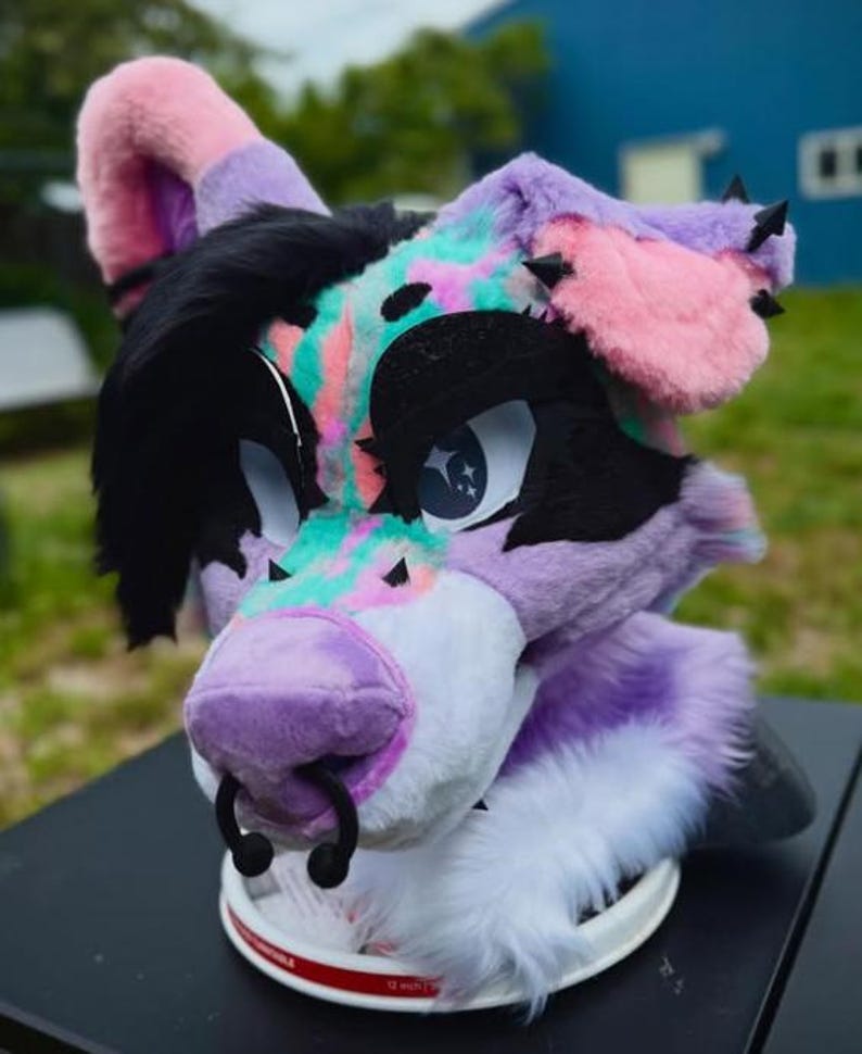 May include: A colorful fursuit head featuring a purple, pink, black, and teal design. The furry head has large, expressive eyes, a purple nose with a black ring, and black spikes on the ears. The fursuit is resting on a white surface.