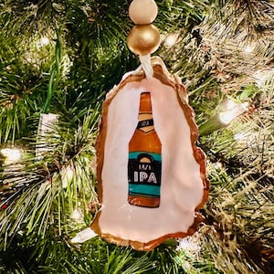May include: Hand-painted oyster shell Christmas ornament featuring a beer bottle labeled "Hazy IPA." The ornament is suspended by a white string with wooden beads and a gold bead. The shell has a natural, textured edge.