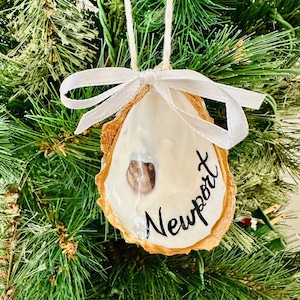 May include: A handcrafted oyster shell Christmas ornament. The shell is painted white with the word "Newport" in black script. A small, brown stone is embedded in the shell. A white ribbon and twine are attached for hanging.