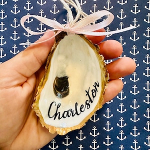 May include: Hand-painted oyster shell ornament with the word "Charleston" in black script. The shell has a gold edge, white interior, and a small dark accent. It is tied with a pink ribbon and string, against a navy blue anchor pattern.