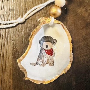 May include: Hand-painted oyster shell ornament featuring a seated dog wearing a red bandana and a dark hat. The shell has a gold-painted edge and is strung with a white cord and wooden beads. A unique, handcrafted item.
