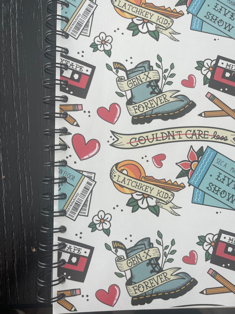 May include: A white notebook with a black spiral binding. The cover features a repeating pattern of colorful illustrations, including hearts, flowers, cassette tapes, boots, and tickets. The text "Latchkey Kid", "Gen-X Forever", "Live Show", and "Couldn't Care Less" is included in the design.