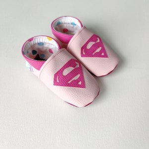 May include: Pink leather baby slippers with a pink Superman logo on the front. The slippers have a colorful polka dot lining.