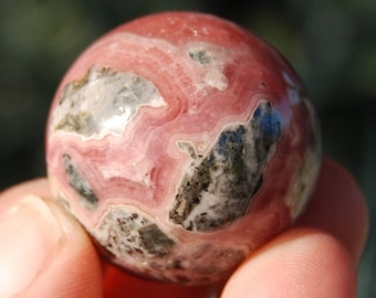 May include: A polished, spherical gemstone with a swirling pattern of pink, gray, and black. The stone is held in a hand, showcasing its smooth, rounded form. The gemstone's colors and patterns create a unique, natural aesthetic.