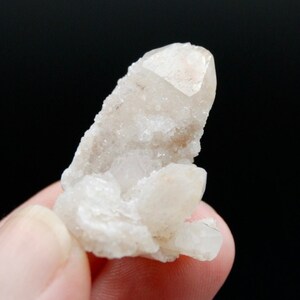 May include: A cluster of small, white quartz crystals with a larger, clear crystal on top. The crystals are clustered together in a natural formation.