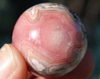 May include: A polished sphere of rhodochrosite, a gemstone with a swirling pattern of pink, white, and gray bands. The sphere is held between fingers, showcasing its smooth, rounded form and natural color variations. The gemstone is approximately 2 inches in diameter.