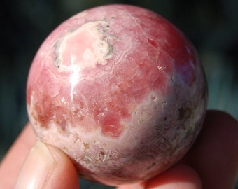 May include: A pink and white rhodochrosite sphere, polished to a smooth finish. The sphere has a swirling pattern of pink and white.