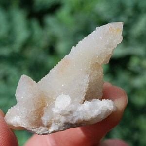 May include: A small, white, crystal cluster with a pointed tip. The crystal has a rough, textured surface and is set on a gray base.