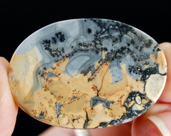 Puede incluir: Una piedra preciosa de forma ovalada con un patrón único. La piedra presenta una mezcla de colores, incluyendo beige, gris y negro, creando un diseño similar a un paisaje. La piedra se sostiene entre los dedos sobre un fondo negro.