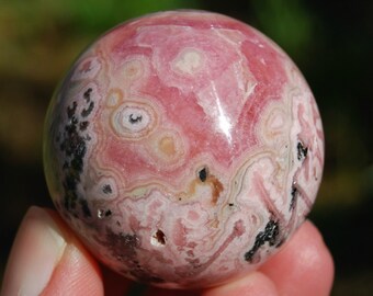 May include: A pink and white sphere of polished pink manganese, also known as rhodochrosite, with black and brown inclusions. The sphere is smooth and has a high gloss finish.