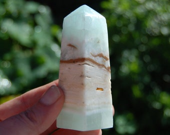 May include: A light blue and white banded aquamarine crystal point, also known as a tower or obelisk, is a polished gemstone. The crystal is a natural stone with brown and white banding.